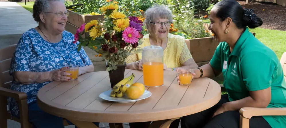 Two residents enjoying a beverage with an employee at an upscale senior living facility at a table outside on a sunny day at The Springs at Willowcreek in Salem, Oregon